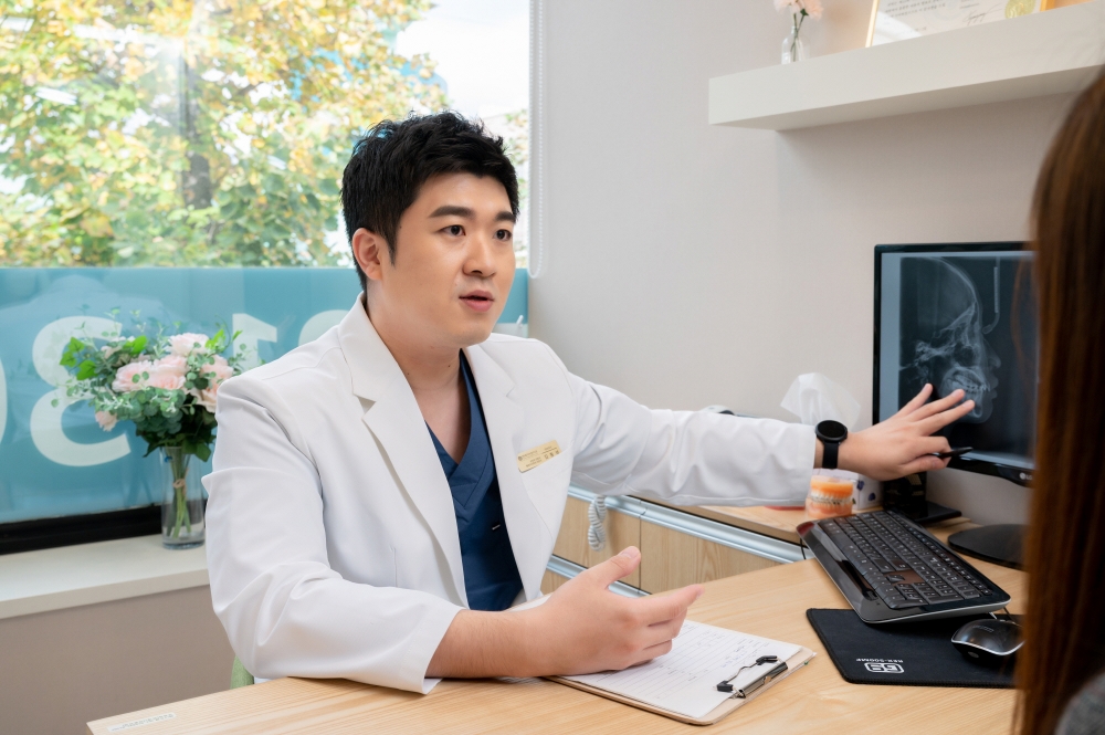 흰 가운 의료진이 상담실에서 모니터에 표시된 측면 두부 방사선 영상을 손으로 가리키며 환자에게 설명하는 장면
